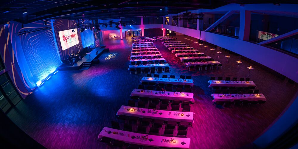 Gro&szlig;er Saal mit langen, parallel angeordneten Tischen und St&uuml;hlen, B&uuml;hne mit beleuchtetem Vorhang und LED Leinwand mit Schriftzug 'Sportlernacht'
