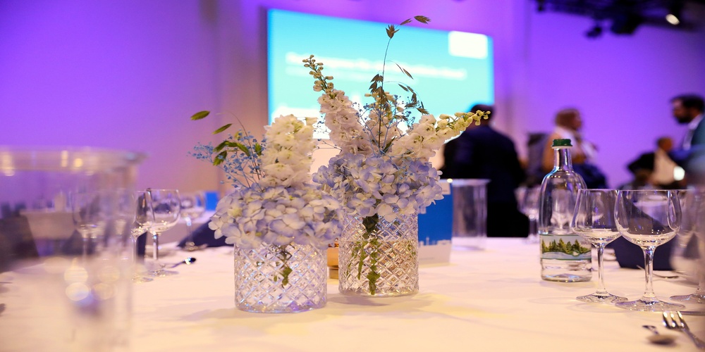 Glasvasen mit wei&szlig;en Blumen auf einem gedeckten Tisch in einem Veranstaltungsraum mit unscharfem Publikum im Hintergrund