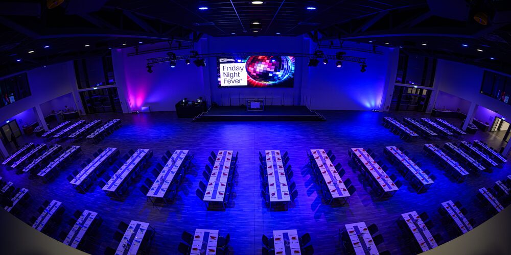 Gro&szlig;er Saal mit Reihen von Tischen und St&uuml;hlen vor einer B&uuml;hne mit LED Leinwand, auf der 'Friday Night Fever' steht, beleuchtet in Blau und Violett.